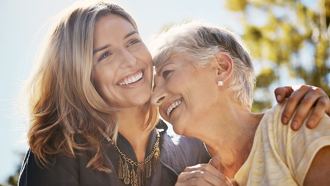 Eine jüngere und eine ältere Frau lächeln und umarmen sich im Freien und genießen gemeinsam einen sonnigen Tag. Die jüngere Frau trägt eine Halskette, die ältere Ohrstecker und ein gestreiftes Hemd. Beide sehen glücklich und zufrieden aus.
