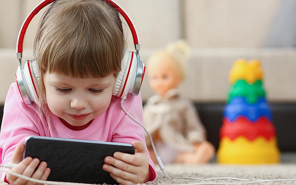 Ein kleines Kind mit großen Kopfhörern liegt auf dem Boden und schaut auf ein Tablet. Im Hintergrund sind eine Puppe und ein buntes Stapelspielzeug zu sehen.