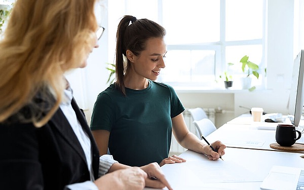 Zwei Frauen an einem Schreibtisch in einem hell erleuchteten Büro. Eine trägt einen schwarzen Blazer und beobachtet, während die andere in einem grünen Hemd ein Dokument unterschreibt. Im Hintergrund sind ein Computer, Pflanzen und eine Tasse zu sehen.