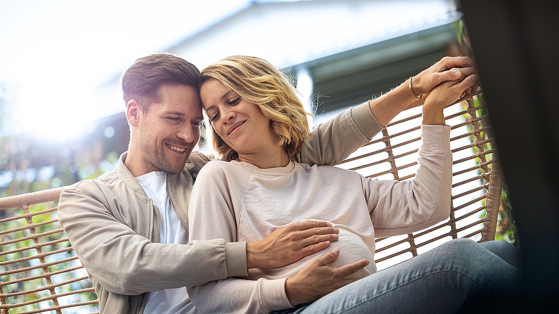Ein lächelndes Paar sitzt draußen auf einer Schaukel. Der Mann schlingt seine Arme um die Frau und legt sanft seine Hände auf ihren schwangeren Bauch. Sie lehnt ihren Kopf an ihn, die Augen geschlossen, und wirkt zufrieden und entspannt. Der Hintergrund ist leicht verschwommen.