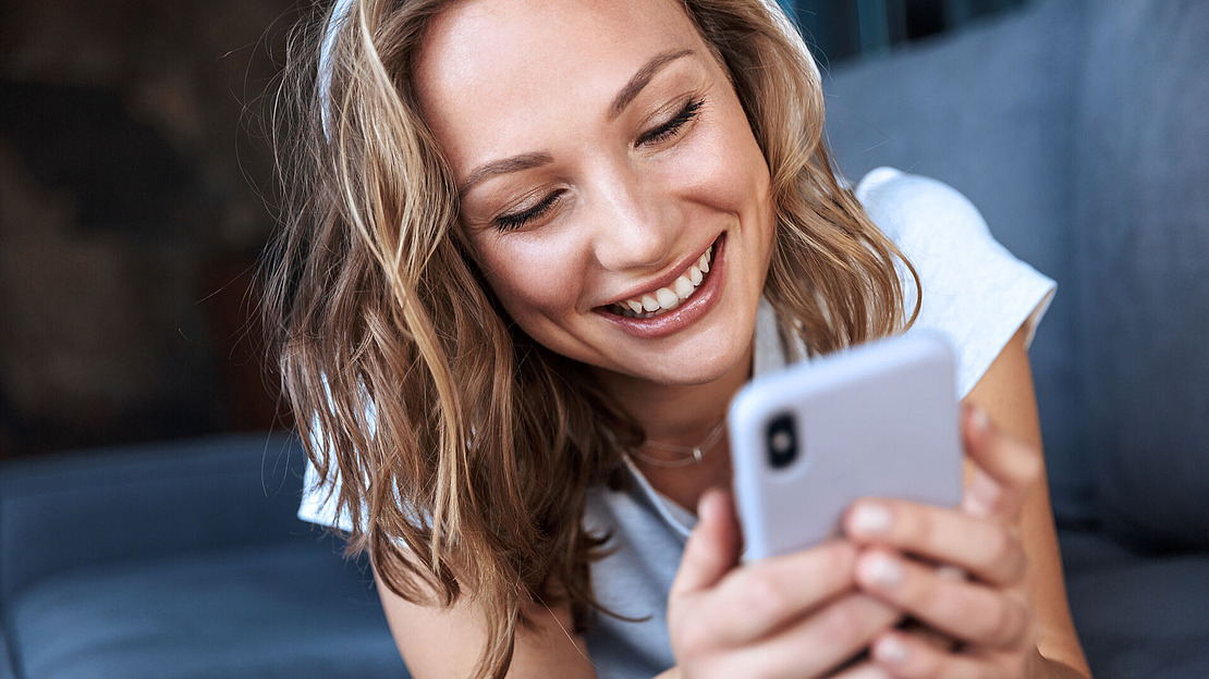 Eine lächelnde Frau mit gewelltem blondem Haar liegt auf einer Couch und hält ein Smartphone in den Händen. Sie scheint glücklich und beschäftigt mit dem, was sie auf dem Bildschirm sieht.