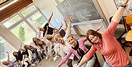 Eine Gruppe lächelnder Frauen sitzt in einem Klassenzimmer und streckt ihre Arme spielerisch diagonal nach außen. Hinter ihnen befinden sich Fenster, eine Tafel mit Schrift und bunte Kunstwerke an der Wand.