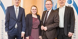 Vier Personen, drei Männer im Anzug und eine Frau im Kleid, stehen lächelnd vor Bannern mit dem Wort „Krankenkasse“ und abstrakten Logos bei einer scheinbar beruflichen oder geschäftlichen Veranstaltung.