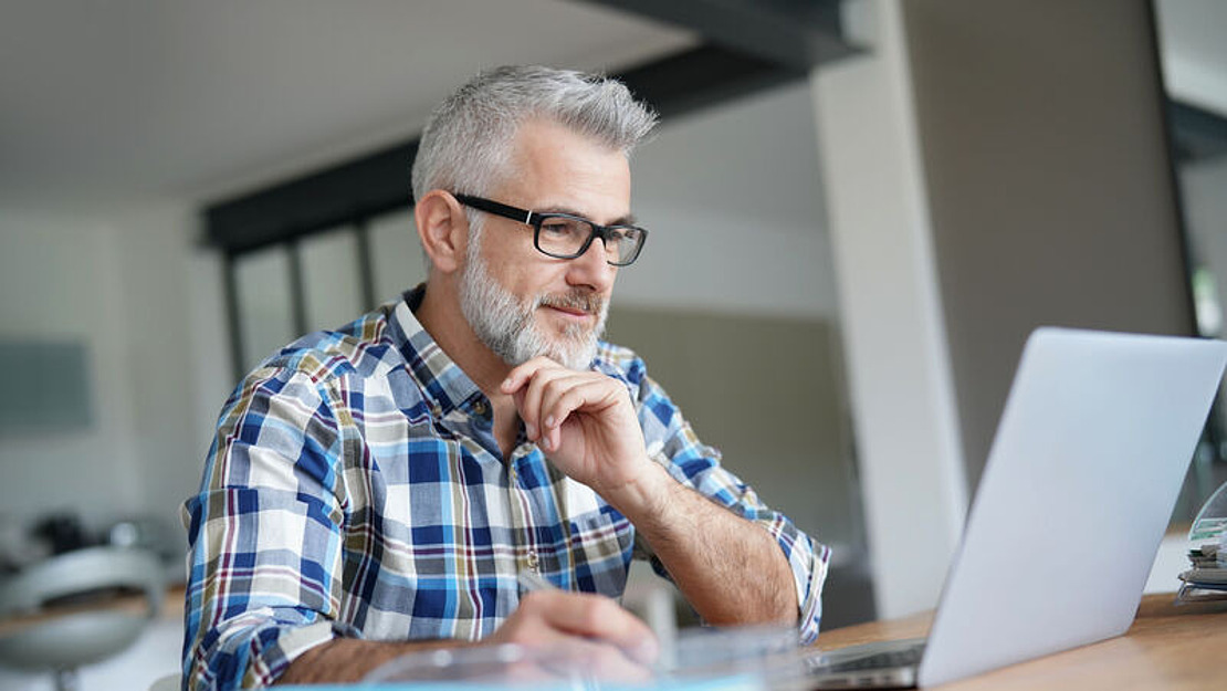Ein Mann mit grauen Haaren und Brille, der ein kariertes Hemd trägt, sitzt an einem Holztisch und arbeitet an einem Laptop. Er stützt sein Kinn auf seine Hand und sieht konzentriert aus. Im Hintergrund ist ein modernes, verschwommenes Interieur zu sehen.