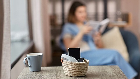 Ein kleiner Korb mit Smartphones und einer TV-Fernbedienung steht auf einem Holztisch neben einer Tasse, während eine Frau im Hintergrund auf einer Couch ein Buch liest.