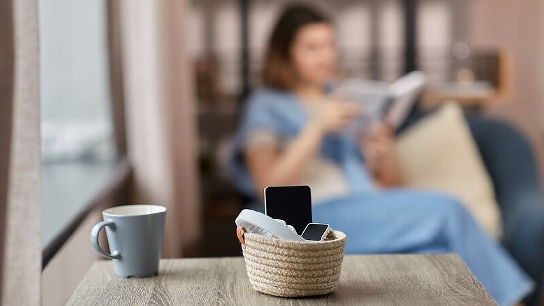 Ein kleiner Korb mit Smartphones und einer TV-Fernbedienung steht auf einem Holztisch neben einer Tasse, während eine Frau im Hintergrund auf einer Couch ein Buch liest.