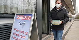 Ein Mann mit Maske und einem Essensbehälter in der Hand verlässt ein Gebäude. Neben ihm hängt ein Schild mit der Aufschrift: „Hier gibt es Essen zum Mitnehmen“.