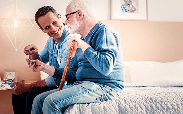 Ein jüngerer und ein älterer Mann sitzen zusammen auf einem Bett, lächeln und betrachten Fotos. Der ältere Mann hält einen Gehstock in der Hand, und sie scheinen einen glücklichen, herzlichen Moment in einem gemütlichen Schlafzimmer zu teilen.