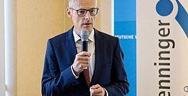 Ein Mann in marineblauem Anzug und Brille spricht bei einer Indoor-Veranstaltung in ein Mikrofon. Er steht vor einem blau-weißen Banner, auf dem teilweise Text zu sehen ist.