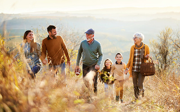 Eine Familie mit mehreren Generationen geht an einem sonnigen Tag gemeinsam durch ein Feld. Sie wirken fröhlich, die Erwachsenen halten sich an den Händen und zwischen ihnen sind zwei Kinder. Die Landschaft ist von sanften Hügeln und herbstlichen Bäumen im Hintergrund geprägt.