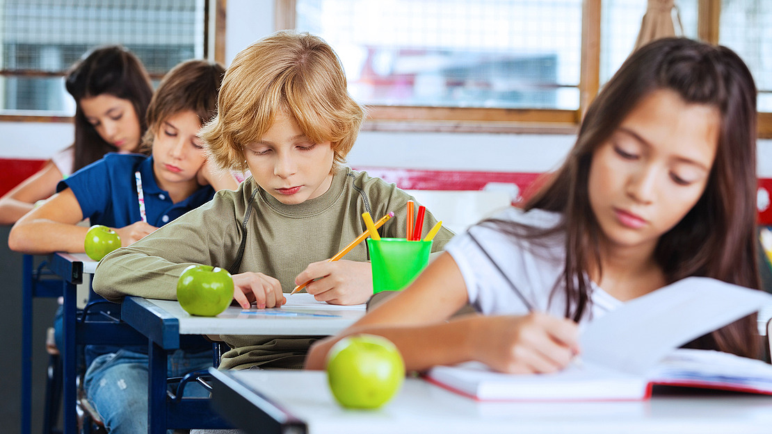 Eine Gruppe von Kindern sitzt an Tischen in einem Klassenzimmer und konzentriert sich auf Schreiben oder Lesen. Auf jedem Tisch liegen grüne Äpfel, und Schulmaterialien sind sichtbar. Das Klassenzimmer ist hell beleuchtet und hat große Fenster im Hintergrund.