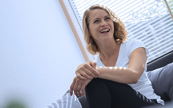 Eine Frau mit schulterlangem blondem Haar sitzt lächelnd auf einem Sofa und blickt leicht nach oben. Sie trägt ein weißes Hemd und eine schwarze Hose. Sonnenlicht fällt durch die Jalousien hinter ihr und erhellt den Raum.
