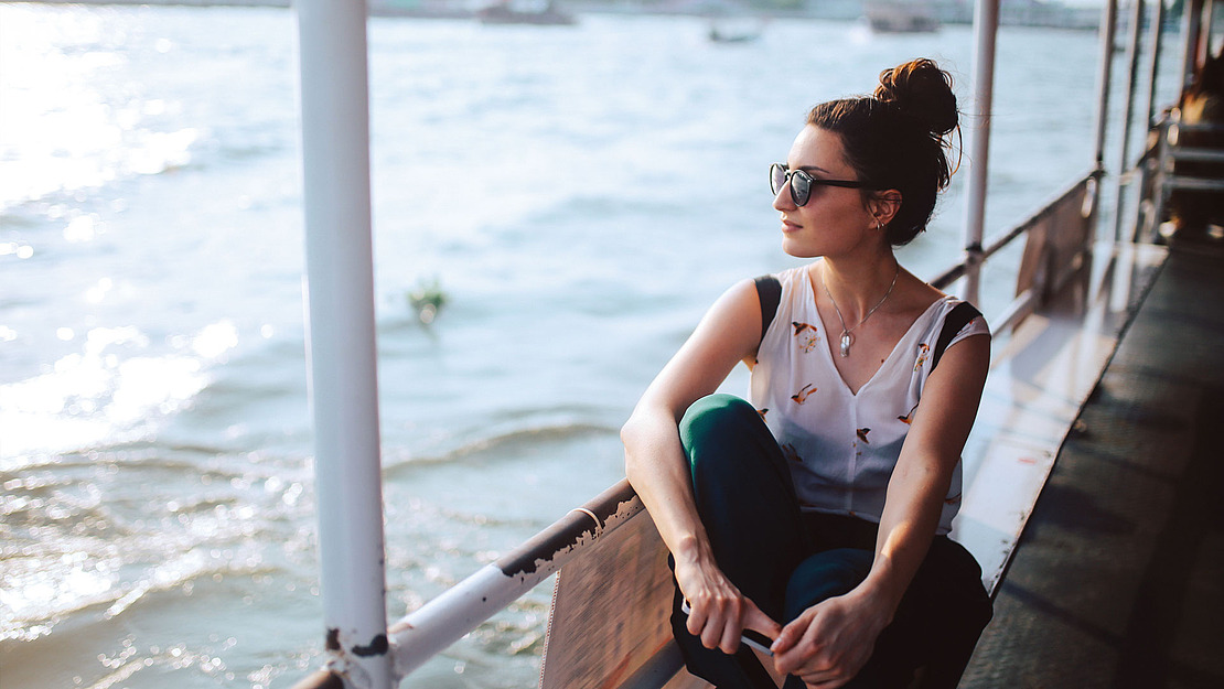 Eine Frau mit Sonnenbrille sitzt auf einem Boot und blickt entspannt auf das Wasser hinaus. Das Sonnenlicht spiegelt sich auf dem Wasser und schafft eine ruhige und friedliche Atmosphäre.