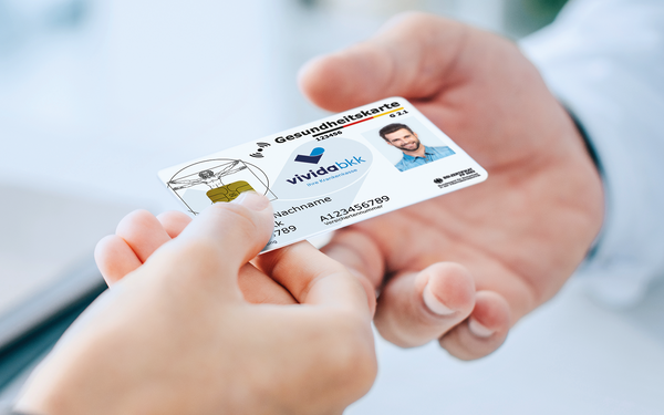 Eine Person überreicht einer anderen Person eine deutsche Krankenversicherungskarte mit Foto und Chip, beide Hände sind in Nahaufnahme zu sehen.