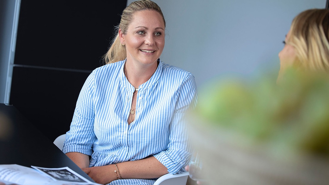 Eine Frau in einem blau-weiß gestreiften Hemd lächelt, während sie an einem Tisch sitzt und Papiere in der Hand hält. Eine andere Person ist teilweise im Vordergrund zu sehen, unscharf, und auch die grünen Pflanzen im Vordergrund sind unscharf.