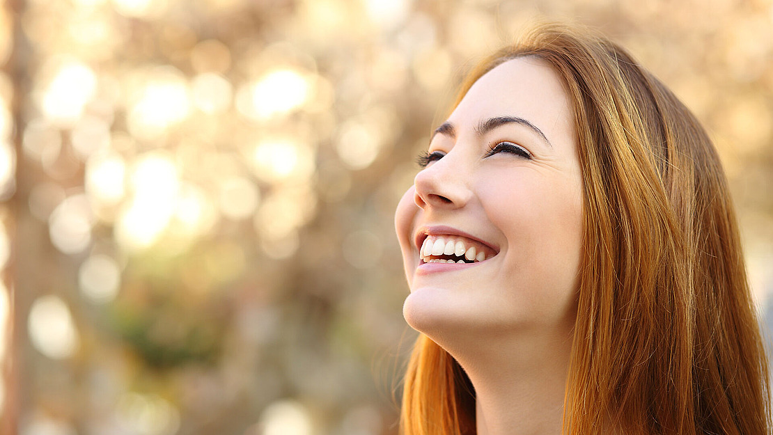 Eine junge Frau mit langem braunem Haar lächelt fröhlich, während sie nach oben blickt, vor dem weichen, verschwommenen Hintergrund warmen Außenlichts.
