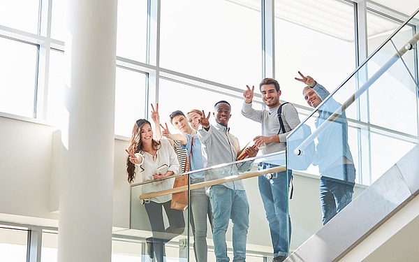 Eine Gruppe von sechs lächelnden Menschen steht auf einem verglasten Balkon im Innenbereich, winkt und macht Peace-Zeichen. Sie tragen Freizeitkleidung, und durch die großen Fenster hinter ihnen strömt Tageslicht.
