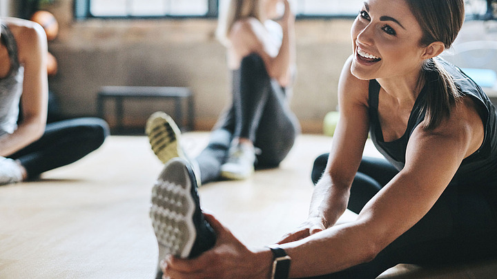 Eine lächelnde Frau in Sportkleidung streckt sich auf dem Boden einer Turnhalle und streckt ihren Fuß nach oben. Im Hintergrund strecken sich andere Menschen. Die Turnhalle ist hell und erhält durch die Fenster natürliches Licht.