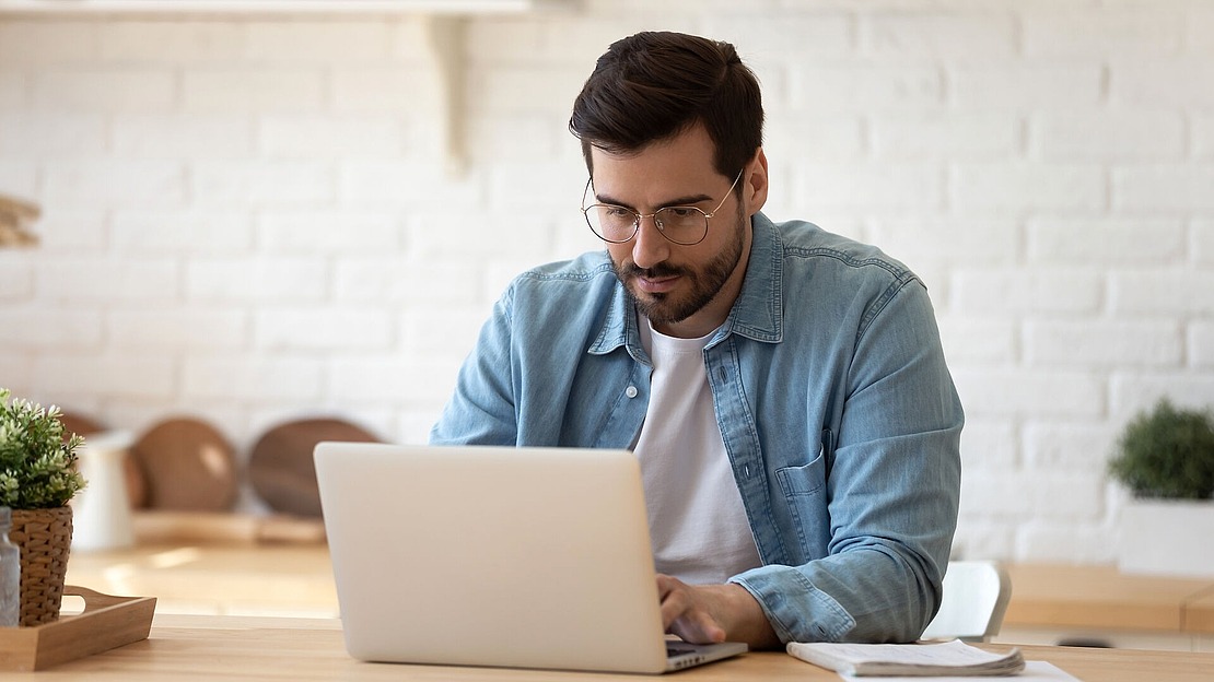 Ein Mann mit dunklem Haar und Brille, der ein helles Jeanshemd über einem weißen T-Shirt trägt, sitzt an einem Tisch und arbeitet an einem Laptop in einem hellen Raum mit weißen Backsteinwänden.