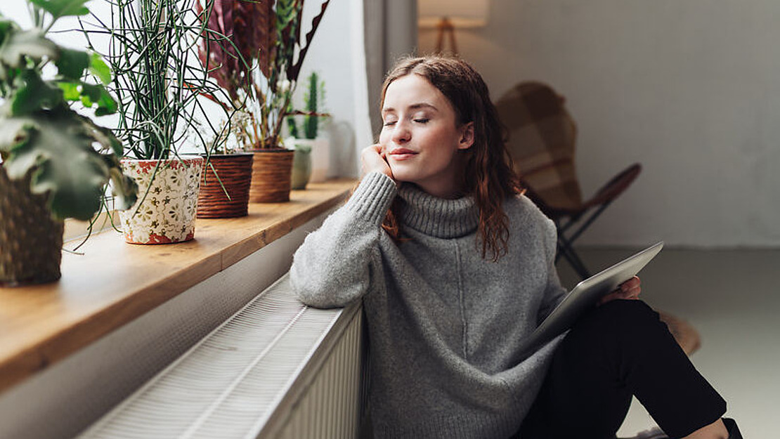 Eine junge Frau in einem grauen Pullover sitzt an einem mit Topfpflanzen gesäumten Fenster, hält ein Tablet in der Hand, lächelt sanft mit geschlossenen Augen und wirkt entspannt und zufrieden.