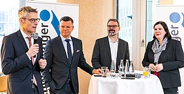 Vier professionell gekleidete Personen stehen mit Wassergläsern und Papieren um einen hohen Tisch, während ein Mann in ein Mikrofon spricht. Im Hintergrund sind zwei Banner mit blauen Logos zu sehen, und große Fenster lassen Tageslicht herein.