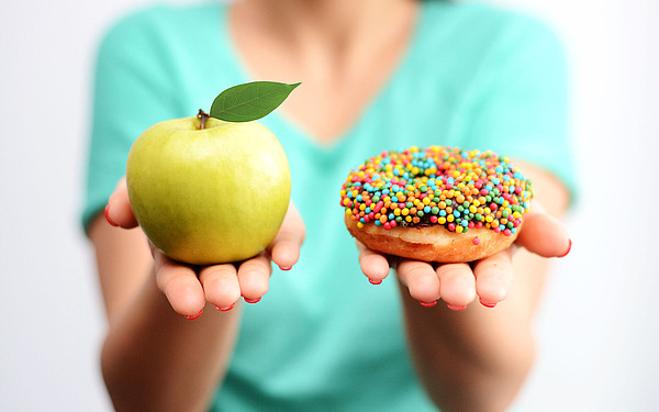 Eine Person in einem blaugrünen Hemd hält in der einen Hand einen grünen Apfel mit Blatt und in der anderen einen mit bunten Streuseln bedeckten Donut und bietet so die Wahl zwischen gesunden und zuckerhaltigen Lebensmitteln.