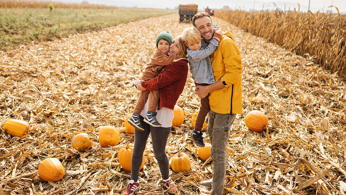 Eine vierköpfige Familie in Herbstkleidung posiert an einem sonnigen Tag glücklich auf einem Kürbisfeld. Die Eltern halten jeweils ein Kind im Arm, umgeben von verstreuten Kürbissen auf einem mit Stroh bedeckten Feld. Im Hintergrund säumen Maisstängel.