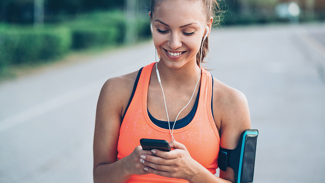 Eine junge Frau in Sportkleidung lächelt und schaut auf ihr Smartphone, während sie Kopfhörer trägt. Sie steht mit einer Telefonarmbinde im Freien und ist bereit für einen Lauf oder ein Training.