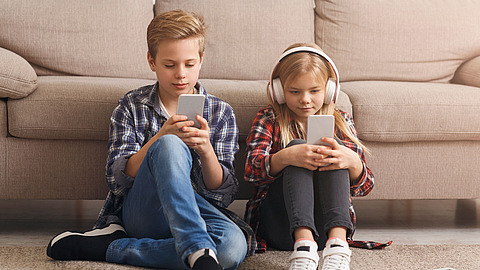 Zwei Kinder sitzen auf dem Boden vor einem Sofa und nutzen jeweils ein Smartphone. Das Mädchen trägt Kopfhörer, und beide Kinder scheinen ganz auf ihre Bildschirme konzentriert zu sein.
