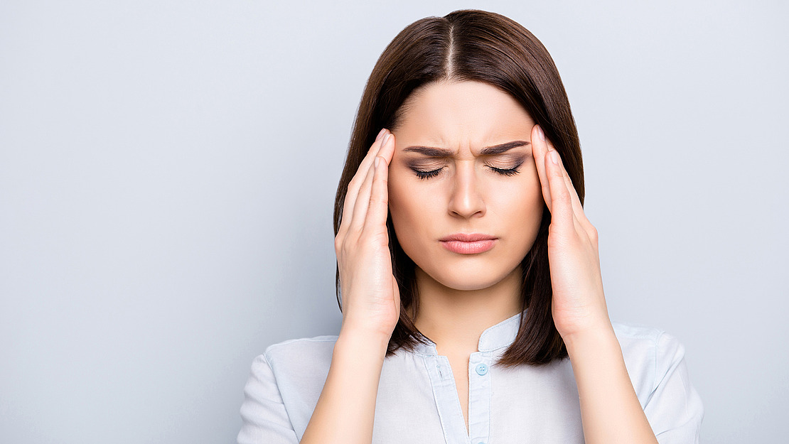 Eine Frau mit schulterlangem braunem Haar schließt die Augen und presst die Finger an die Schläfen. Sie wirkt, als hätte sie Kopfschmerzen oder sei gestresst. Sie trägt eine helle Bluse und steht vor einem schlichten Hintergrund.