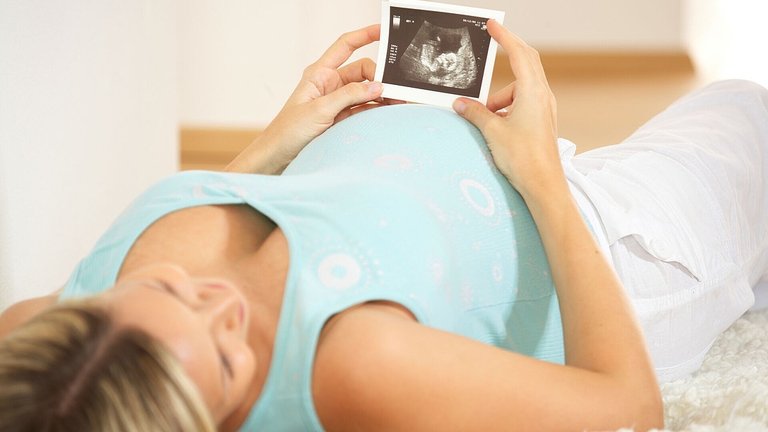 Eine schwangere Frau in einem hellblauen Oberteil liegt auf dem Rücken und hält ein Ultraschallfoto ihres Babys über ihren Bauch und betrachtet es.
