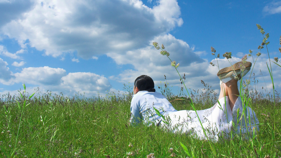 Eine Person in einem weißen Kleid liegt auf dem Bauch in einem grasbewachsenen Feld unter einem blauen Himmel mit vereinzelten Wolken und blickt in die Ferne. Hohes Gras umgibt sie und schafft eine friedliche, sommerliche Szene.