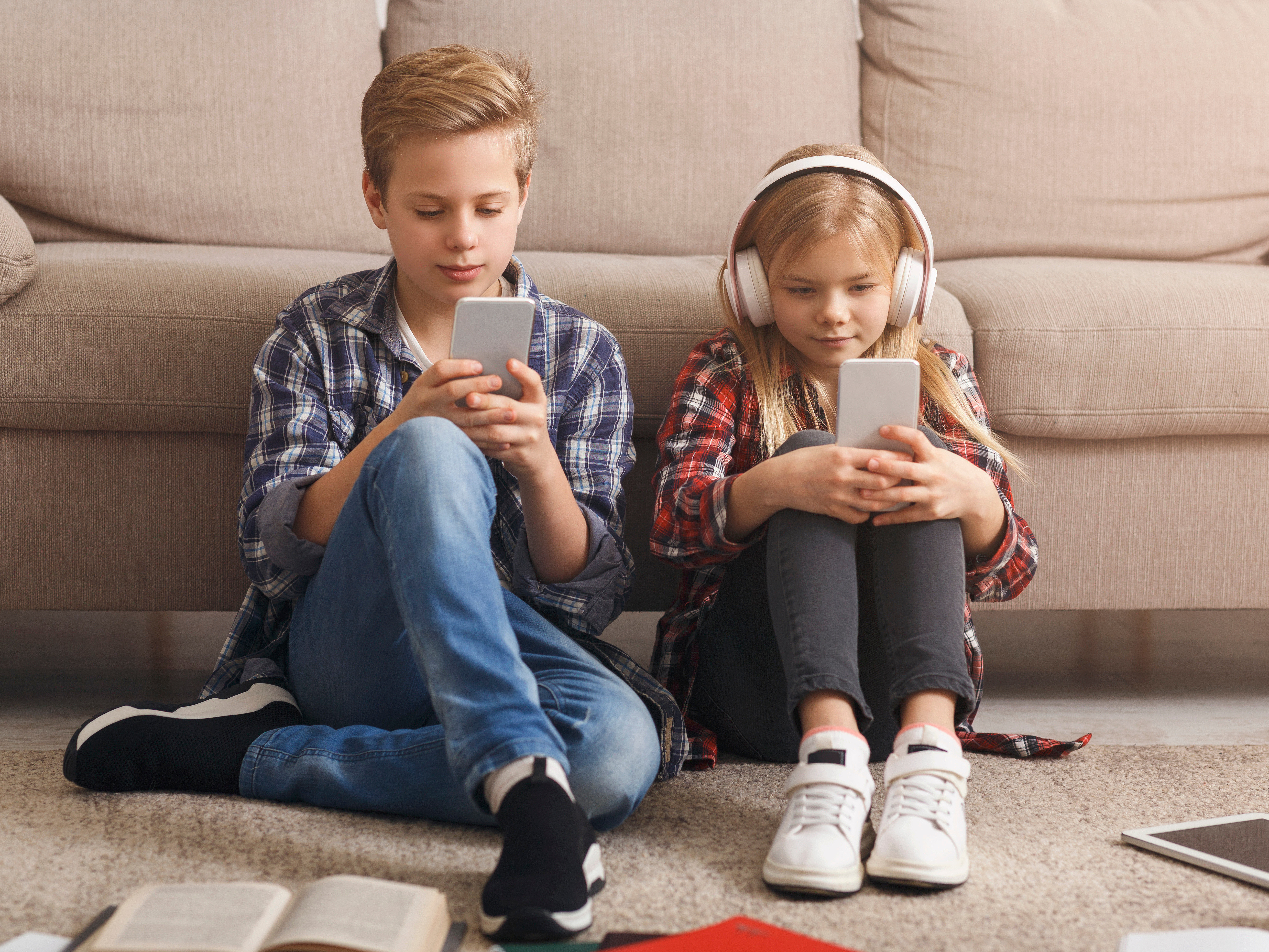 Zwei Kinder sitzen auf einem Teppich vor einem Sofa und konzentrieren sich auf ihre Smartphones. Der Junge trägt ein kariertes Hemd und Jeans, das Mädchen Kopfhörer und ein kariertes Hemd. Bücher und ein Tablet liegen daneben auf dem Boden.
