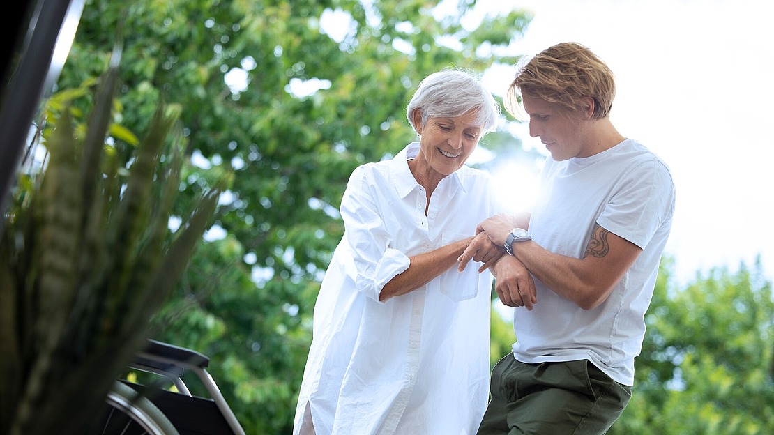 Eine ältere Frau und ein jüngerer Mann, beide lächelnd, stehen im Freien neben einem Rollstuhl. Der Mann stützt die Frau beim Aufstehen, während im Hintergrund Sonnenlicht und grüne Bäume zu sehen sind.