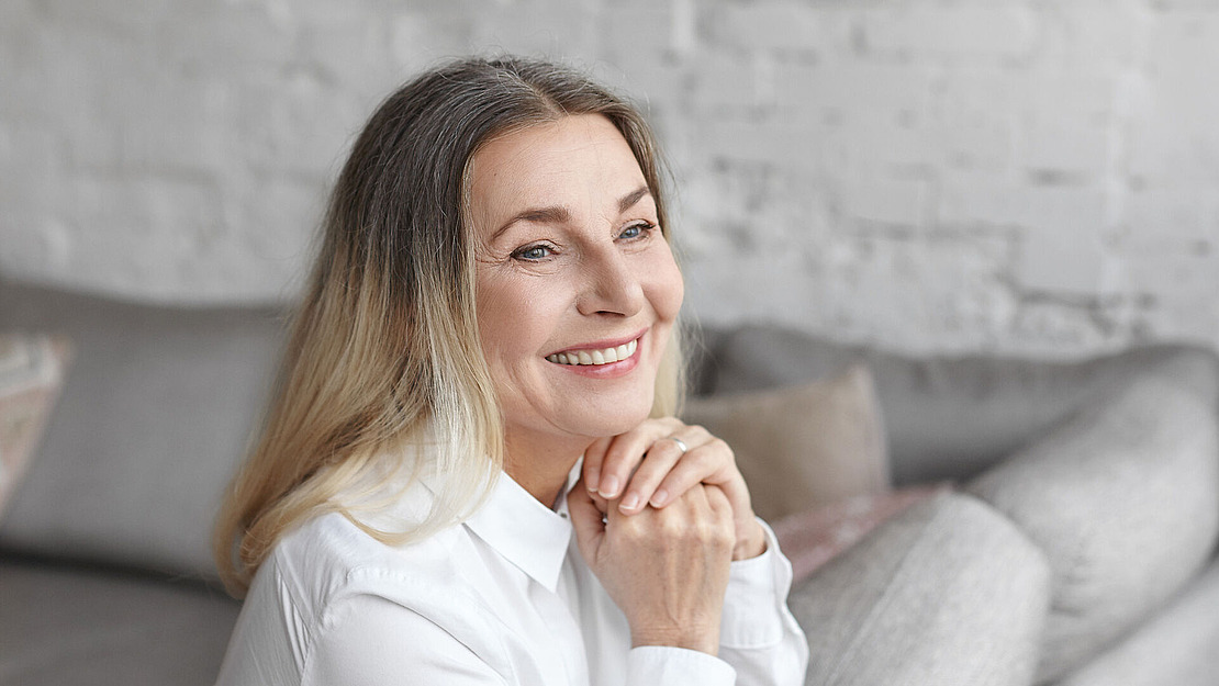 Eine lächelnde ältere Frau mit langem, ergrauendem Haar und einer weißen Bluse sitzt im Haus, die Hände unter dem Kinn verschränkt, und blickt zur Seite. Im Hintergrund sind ein helles Sofa und eine weiße Backsteinwand zu sehen.