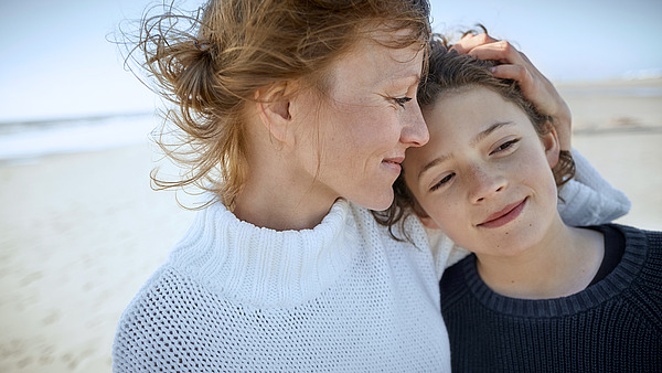 Eine Frau legt ihre Hand sanft auf den Kopf eines Kindes, während beide lächeln. Sie stehen dicht beieinander an einem Strand, im Hintergrund ist das Meer zu sehen. Die Frau trägt einen weißen Pullover und das Kind einen dunklen.