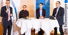Vier Personen in Businesskleidung stehen oder sitzen an zwei runden Tischen mit weißen Tischdecken und sprechen bei einer Veranstaltung. Ein Mann spricht in ein Mikrofon; dahinter sind Transparente mit der Aufschrift „Krankenkasse“ und „Die Schwenninger“ zu sehen.