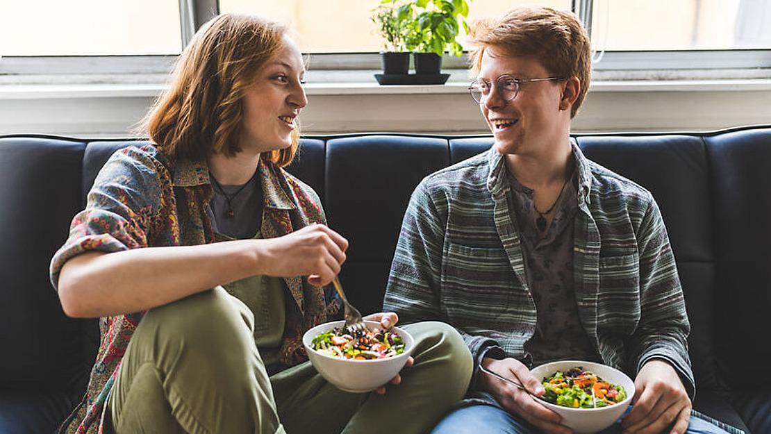 Zwei junge Erwachsene sitzen lächelnd auf einem Sofa und halten Schüsseln mit Salat in den Händen. Auf dem Fensterbrett hinter ihnen steht eine kleine Topfpflanze.