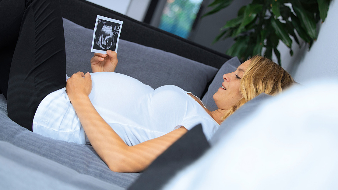 Schwangere Frau liegt lächelnd auf einer Couch und hält ein Ultraschallbild in der Hand. Sie trägt ein weißes Hemd und eine schwarze Hose. Im Hintergrund ist eine grüne Pflanze zu sehen.