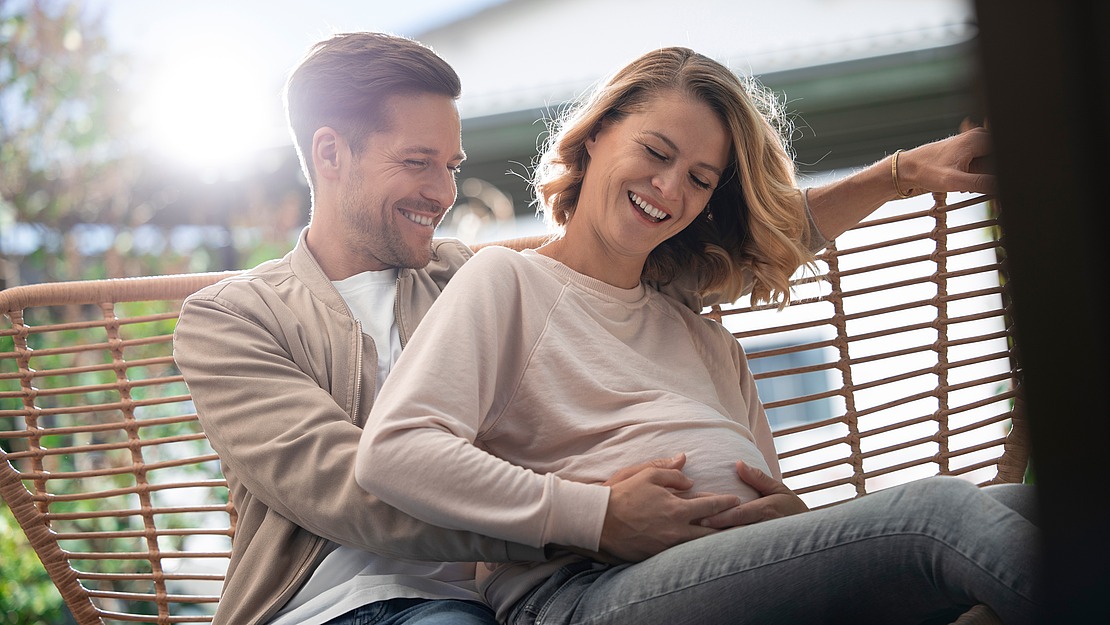Ein lächelndes Paar sitzt draußen auf einer Schaukel. Der Mann umarmt liebevoll die schwangere Frau, die ihre Hände auf ihrem Bauch ruhen lässt. Sie genießen einen sonnigen Tag mit einem Haus und viel Grün im Hintergrund.