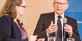 Ein Mann im Anzug spricht in ein Mikrofon und gestikuliert dabei mit der Hand, während eine Frau während einer Diskussion an einem Tisch mit zwei Gläsern Wasser aufmerksam zuhört.