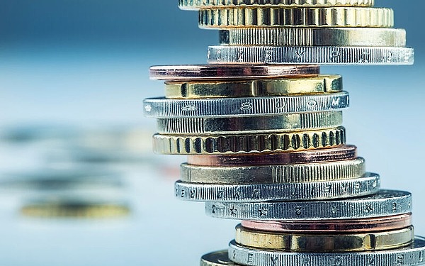 Ein Stapel verschiedener Münzen in Silber-, Gold- und Bronzefarben, ungleichmäßig gestapelt vor einem verschwommenen blauen Hintergrund. Die Münzen haben unterschiedliche Randmuster und Inschriften, wodurch ein strukturiertes Erscheinungsbild entsteht.