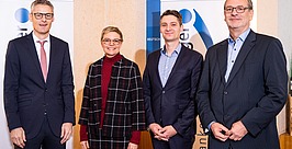 Vier professionell gekleidete Personen, zwei Männer in Anzügen, eine Frau in einem karierten Mantel und einem roten Rollkragenpullover und ein weiterer Mann in einer Nadelstreifenjacke, stehen im Innenbereich vor Bannern mit blau-weißen Mustern.