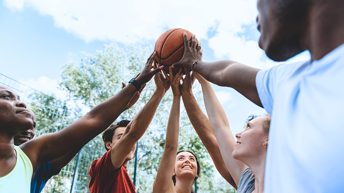 Eine Gruppe junger Erwachsener im Freien, die mit erhobenen Armen in einem Kreis stehen, alle halten einen Basketball zusammen und lächeln, mit Bäumen und einem blauen Himmel im Hintergrund.