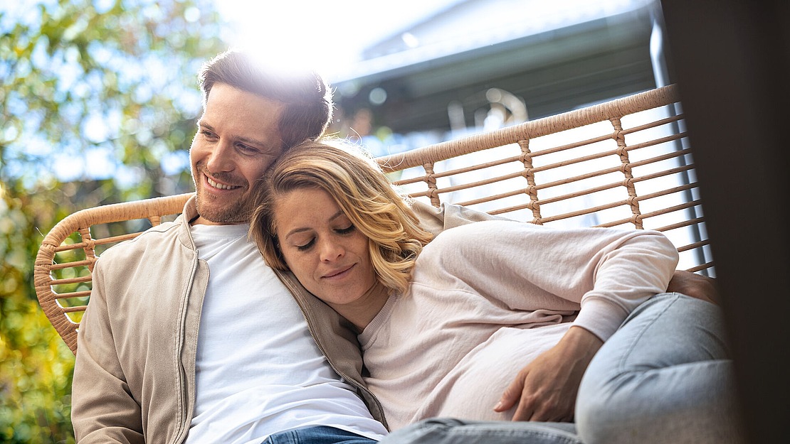 Ein Paar sitzt eng aneinander gekuschelt auf einer Gartenschaukel. Die Frau, die einen leichten Pullover trägt, lehnt mit geschlossenen Augen an der Schulter des Mannes. Der Mann, in einer leichten Jacke, lächelt und blickt in die Ferne. Sonnenlicht fällt durch die Bäume.