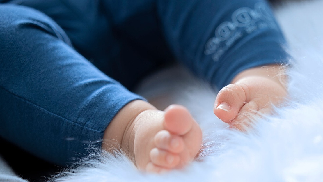 Eine Nahaufnahme der nackten Füße eines Babys, das auf einer weichen, weißen, flauschigen Oberfläche ruht. Das Baby trägt eine blaue Hose, und der Fokus liegt auf den Füßen und Zehen.