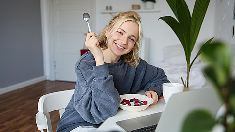 Eine lächelnde Frau sitzt an einem Tisch in einem hellen Raum, in der Hand einen Löffel und eine Schale mit Obst und Joghurt. Eine Kaffeetasse, ein Laptop und eine Grünpflanze stehen ebenfalls auf dem Tisch. Sie wirkt entspannt und fröhlich.