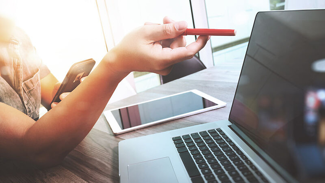 Eine Person hält ein Smartphone in der Hand und zeigt mit einem Rotstift auf einen Laptop-Bildschirm auf einem Holztisch, auf dem auch ein Tablet zu sehen ist. Das Sonnenlicht fällt durch ein Fenster im Hintergrund.