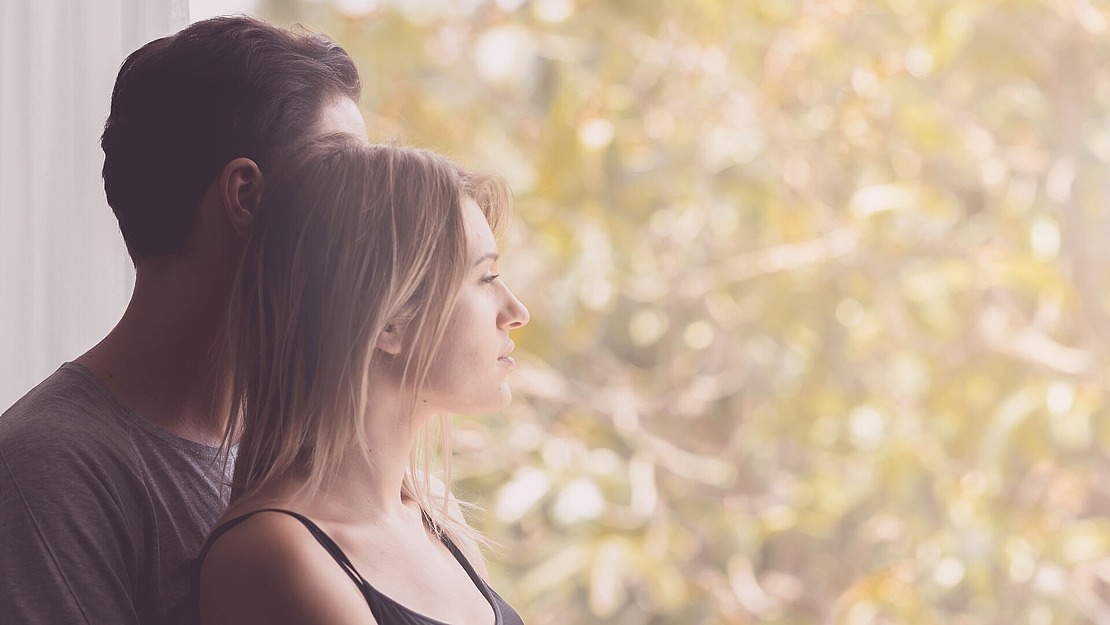 Ein Mann und eine Frau stehen dicht beieinander und blicken aus einem Fenster mit sanftem Sonnenlicht und verschwommenen Grünflächen im Hintergrund. Die Szene hat eine ruhige und kontemplative Stimmung.