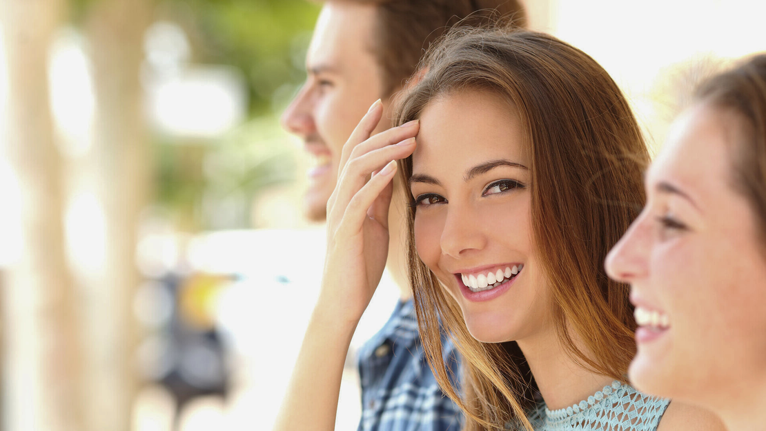 Drei junge Erwachsene lächeln im Freien. Die Frau in der Mitte blickt in die Kamera und berührt ihr Haar, während die beiden anderen neben ihr leicht unscharf sind und im natürlichen Licht fröhlich wirken.
